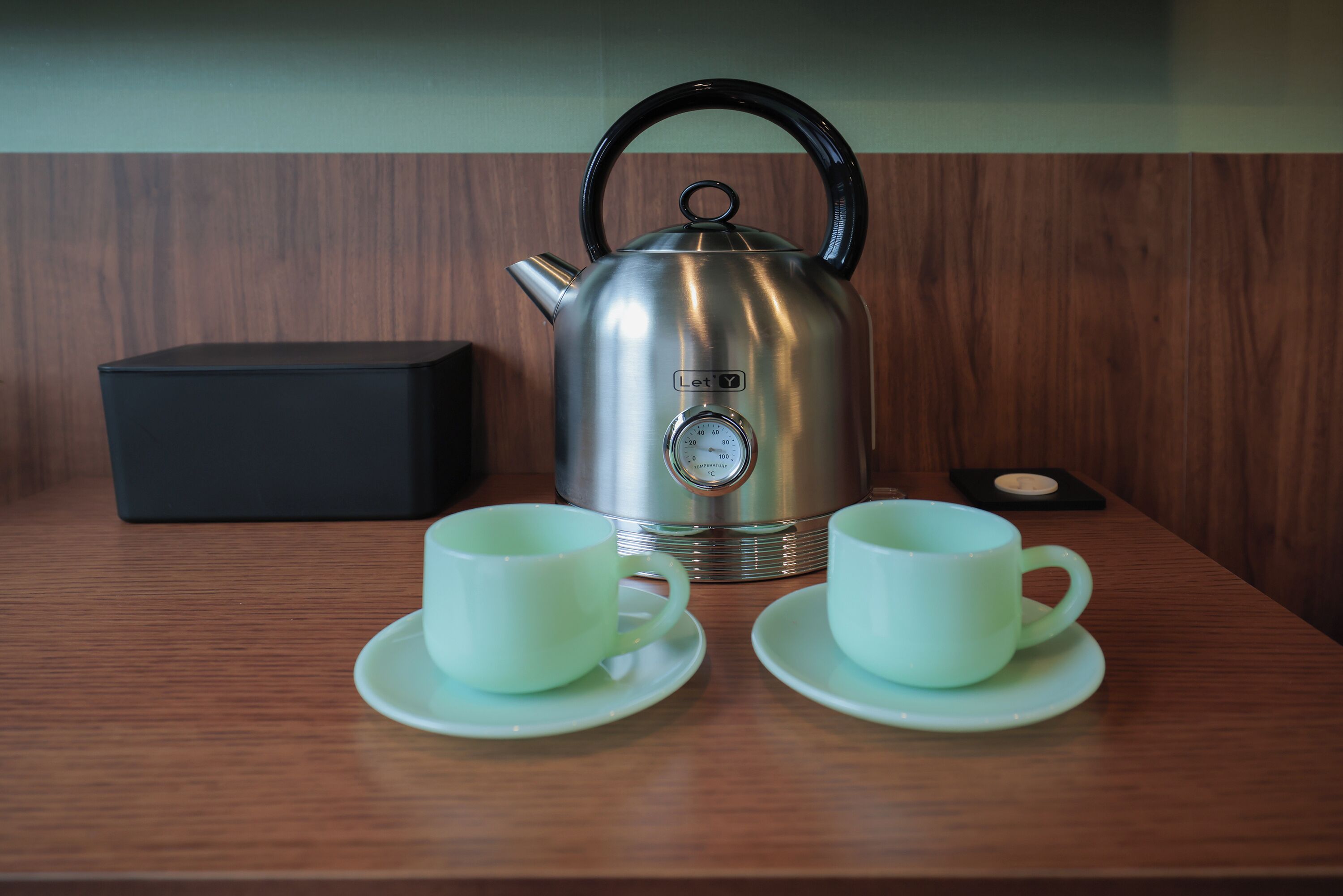 A stainless steel kettle with a black handle, two white cups with saucers, and a black box on a wooden table.