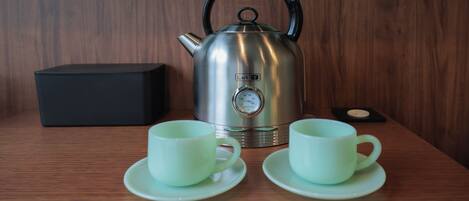 A stainless steel kettle with a black handle, two white cups with saucers, and a black box on a wooden table.