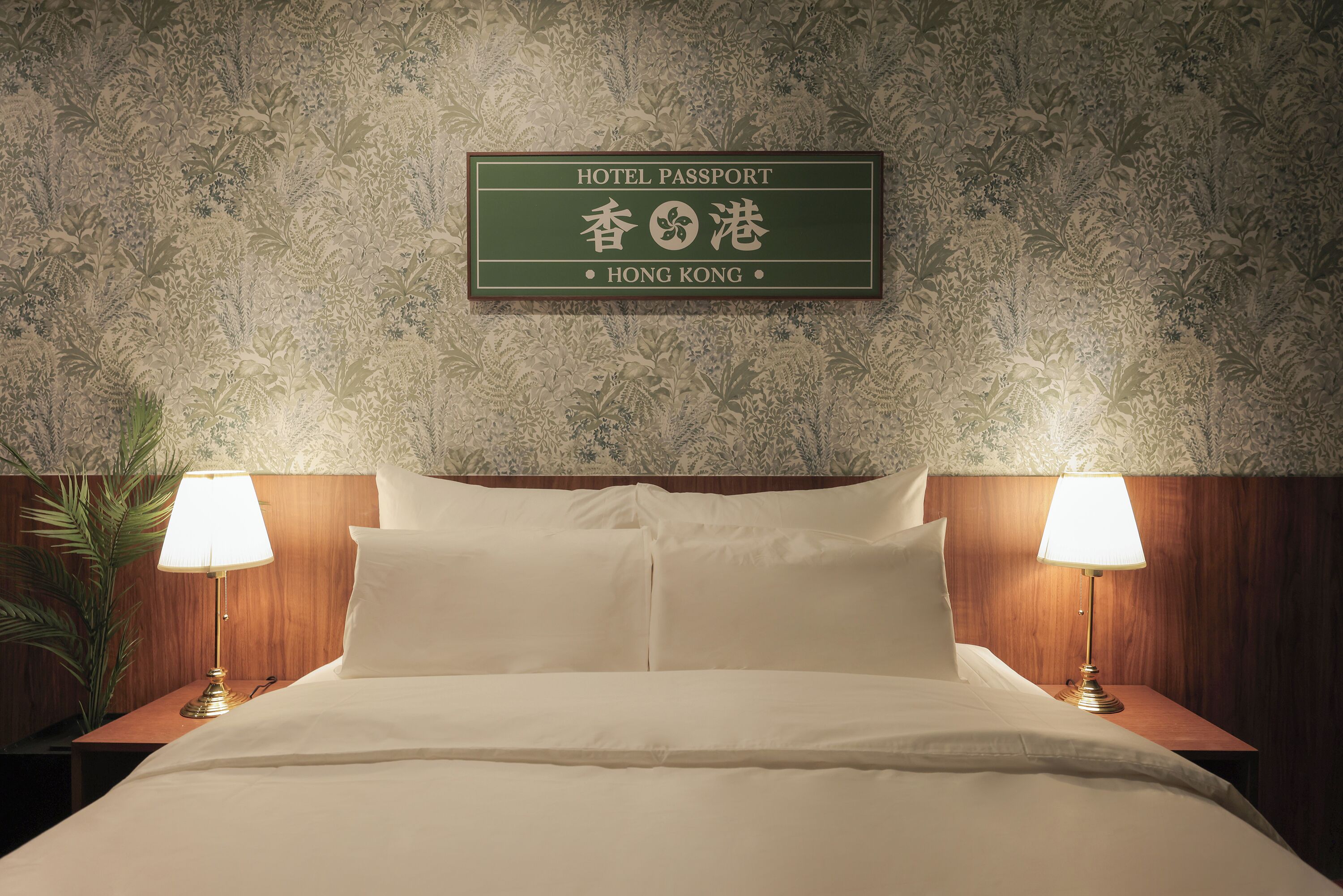 A hotel room with a bed, two bedside lamps, a nightstand, and a wall-mounted sign indicating the hotel is in Hong Kong.