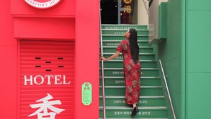 Eine Frau steigt eine grüne Treppe vor einem Gebäude mit einer roten Tür herunter, an der das Wort 'HOTEL' und koreanische Schriftzeichen stehen.