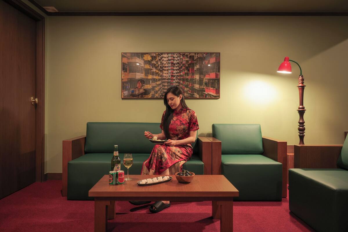 A woman sitting on a green sofa in a room with a wooden table, a red lamp, and a painting on the wall.