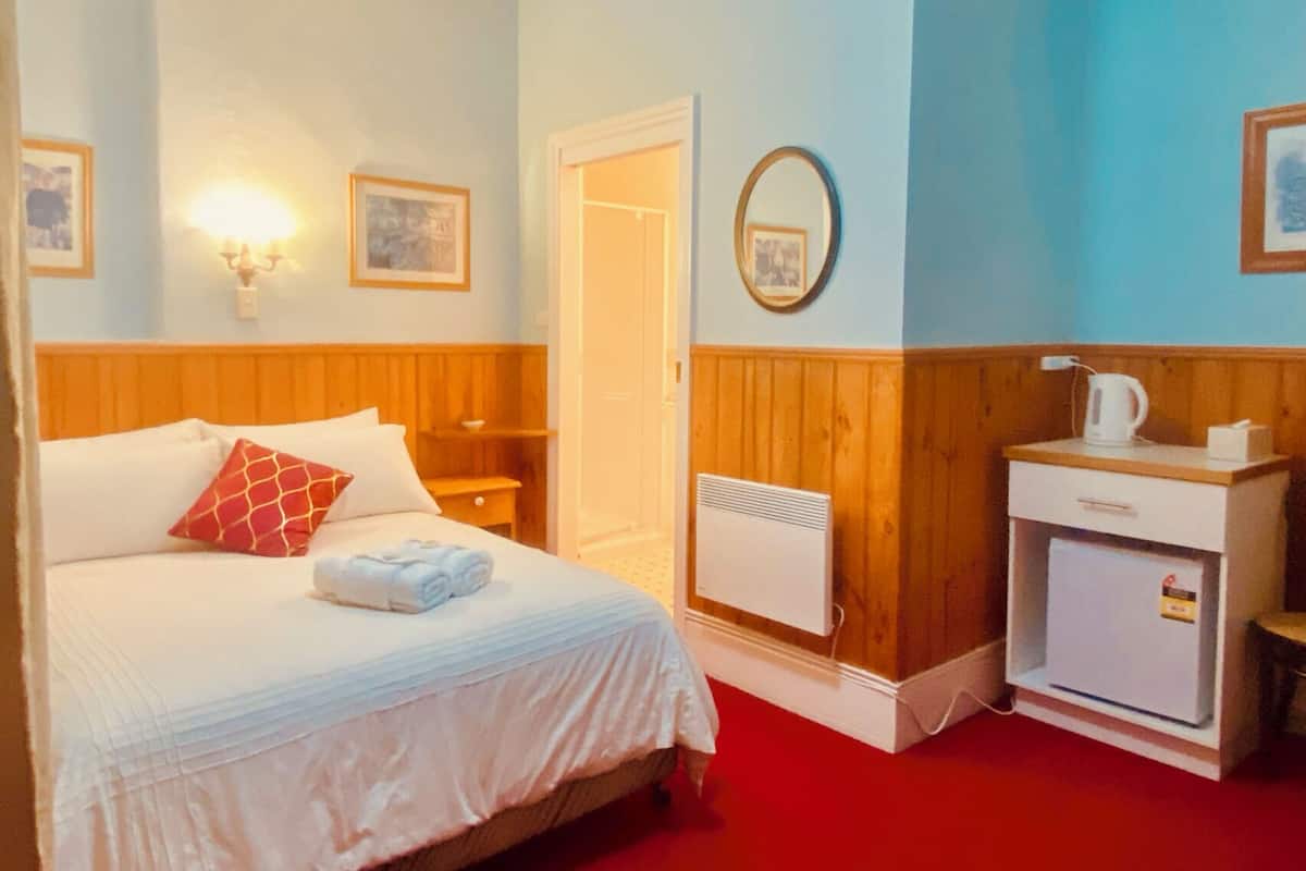 Quarto de hotel com cama, mesa de cabeceira, frigorífico branco pequeno e espelho fixado na parede.
