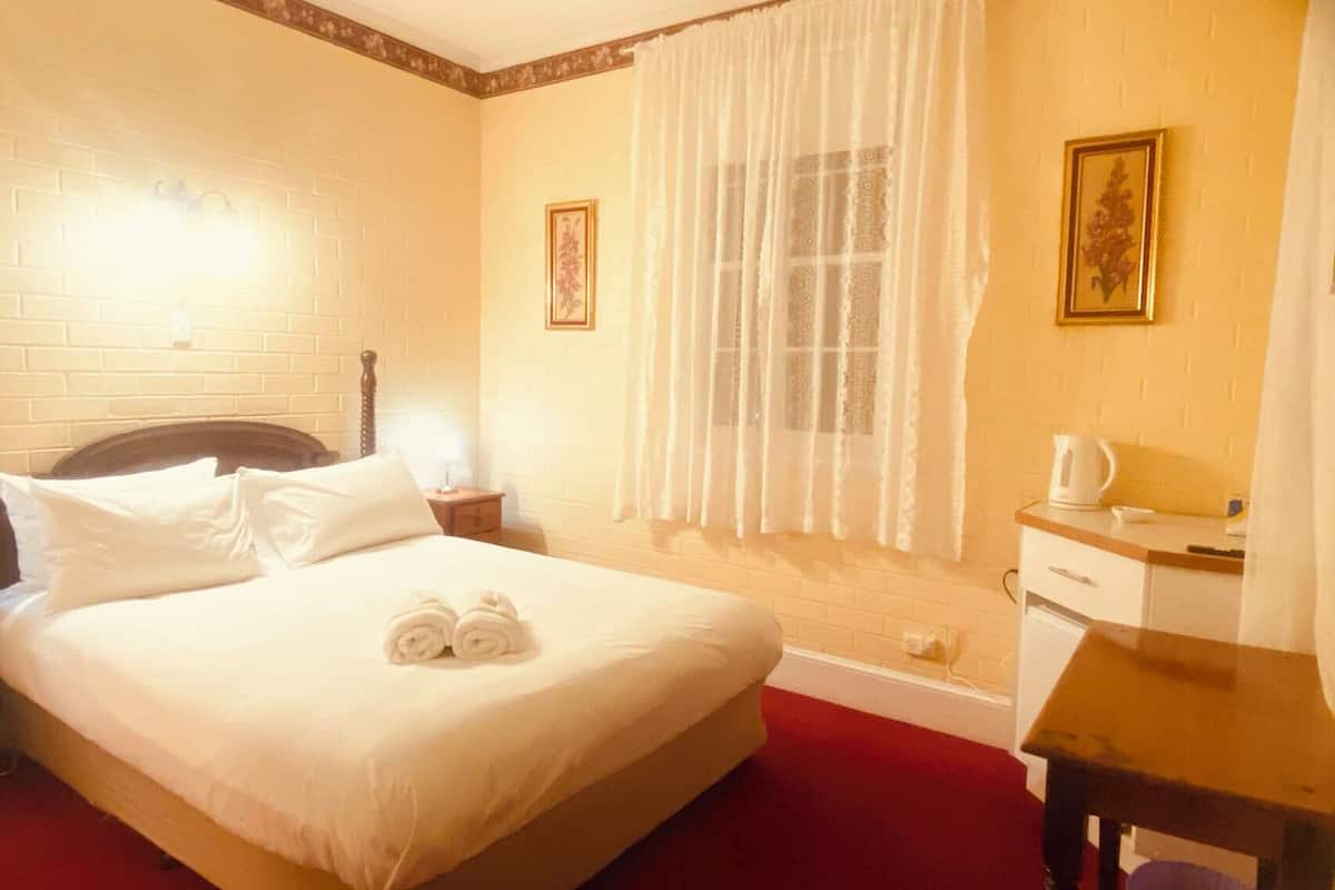 Quarto de hotel com cama, mesa de cabeceira, escrivaninha, janela com cortinas e duas imagens emolduradas na parede.