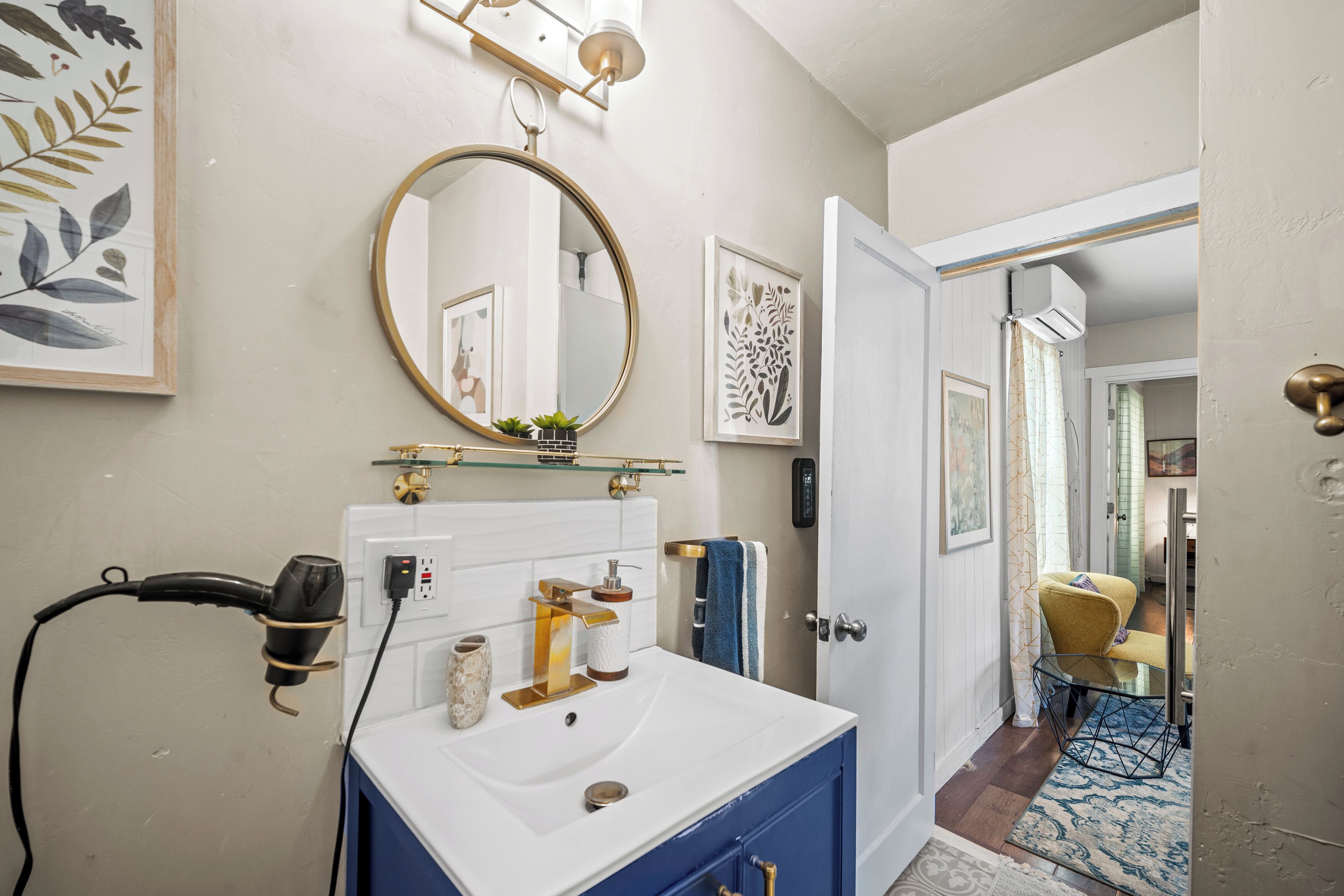 Une salle de bain avec un lavabo blanc, un robinet doré et un miroir rond. Un sèche-cheveux est branché, et une petite plante est posée sur le lavabo.