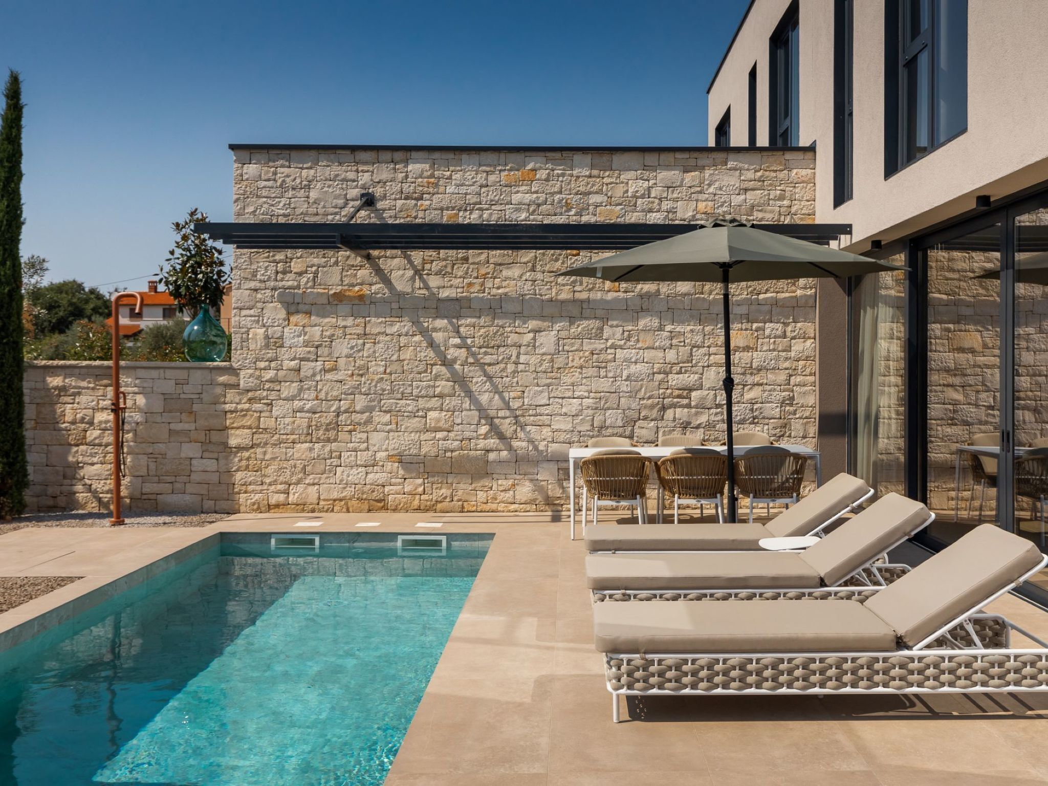 Un espace piscine extérieur moderne, agrémenté d’un mur en pierre, de chaises longues et d’une table avec des parasols.