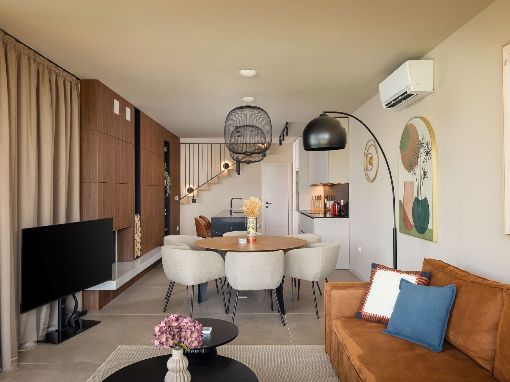 Ein modernes Wohnzimmer mit Flachbildfernseher, einem runden Esstisch mit Stühlen, einer Couch mit Kissen, einem Couchtisch mit Blumen und einer Treppe, die zu einer Küchenzeile führt.