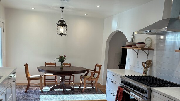 A modern kitchen with wooden flooring, a dining table with chairs, a stove, and a hanging light fixture.