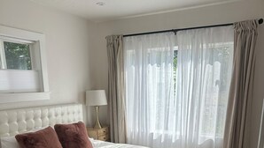A neatly arranged bedroom with a large bed, a bedside table with a lamp, and sheer curtains on the window.