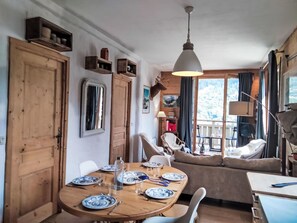 Ein Essbereich mit einem Holztisch, gedeckt für vier Personen, umgeben von weißen Stühlen. Eine Couch mit Kissen, eine Lampe und ein Blick ins Freie durch ein großes Fenster.