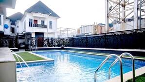 Una piscina con escalera metálica, un edificio blanco con balcón y un muro decorativo con diseño geométrico.