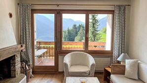 A cozy room with a wooden floor, a white sofa, a chair, a small table with a glass, and a fireplace. Large windows offer a view of mountains and greenery. - Toissa A4 by Interhome (Savognin)