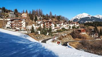 Un village de montagne enneigé, avec des bâtiments à plusieurs étages, une route sinueuse et un ciel bleu dégagé.