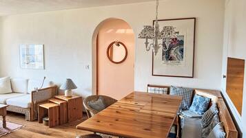 Un salon chaleureux avec une table à manger en bois, des meubles en osier, un canapé blanc et, au mur, un tableau encadré provenant d’une galerie d’art.