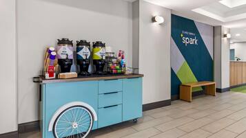 A blue cart with beverage dispensers, a wooden bench, and a wall with a geometric design and the text 'It only takes a spark.'