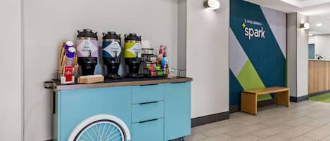 Un chariot bleu équipé de distributeurs de boissons, un banc en bois et un mur orné d’un motif géométrique ainsi que de l’inscription : « Il suffit d’une étincelle. »