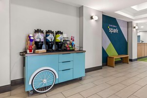 Un carrito azul con dispensadores de bebidas, un banco de madera y una pared con diseño geométrico y el texto 'It only takes a spark'.