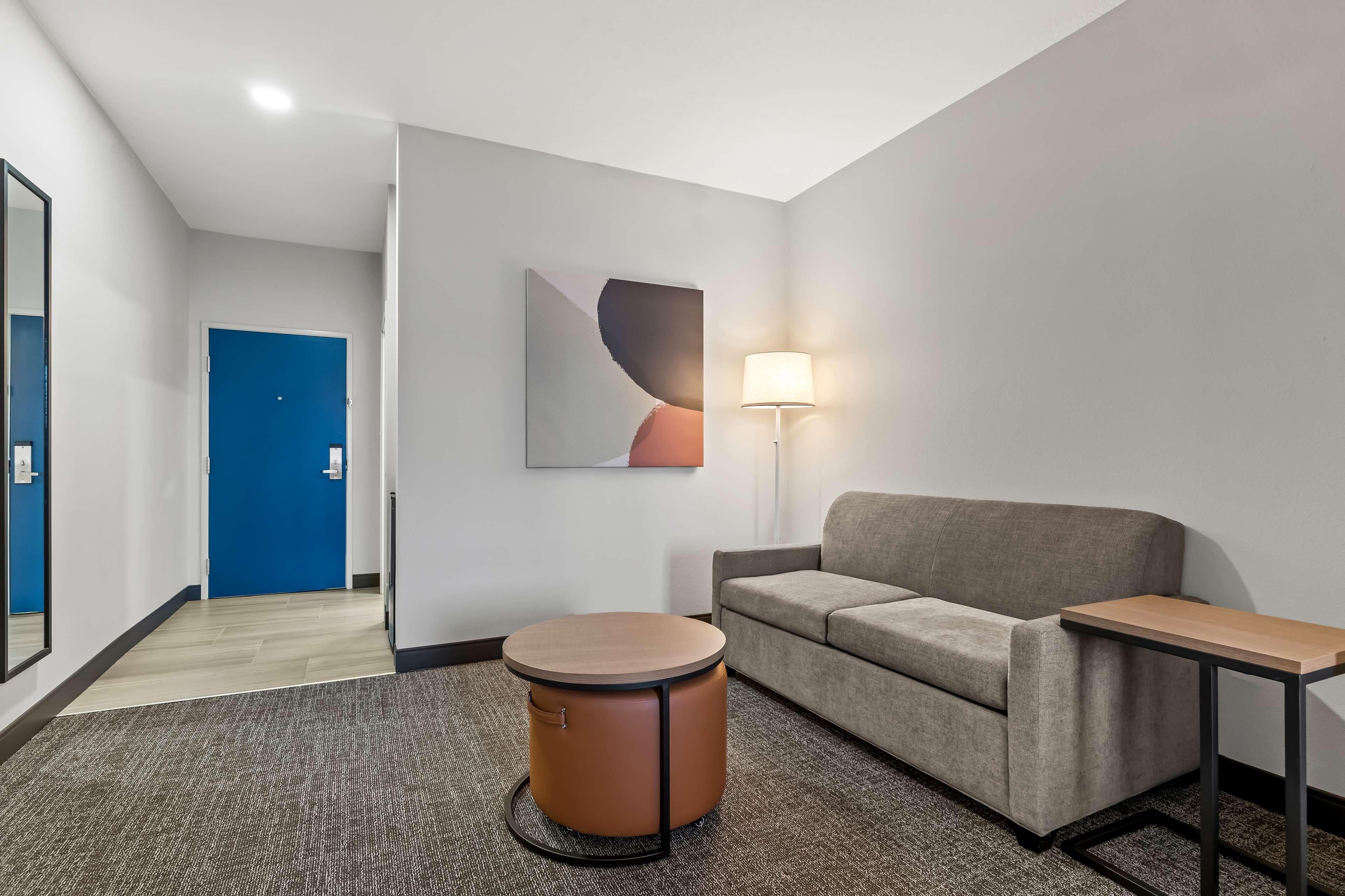 A modern hotel room with a grey sofa, a round wooden side table, a round wooden stool, a lamp, and a blue door.