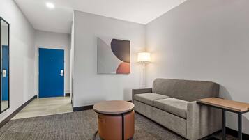 A modern hotel room with a grey sofa, a round wooden side table, a round wooden stool, a lamp, and a blue door.
