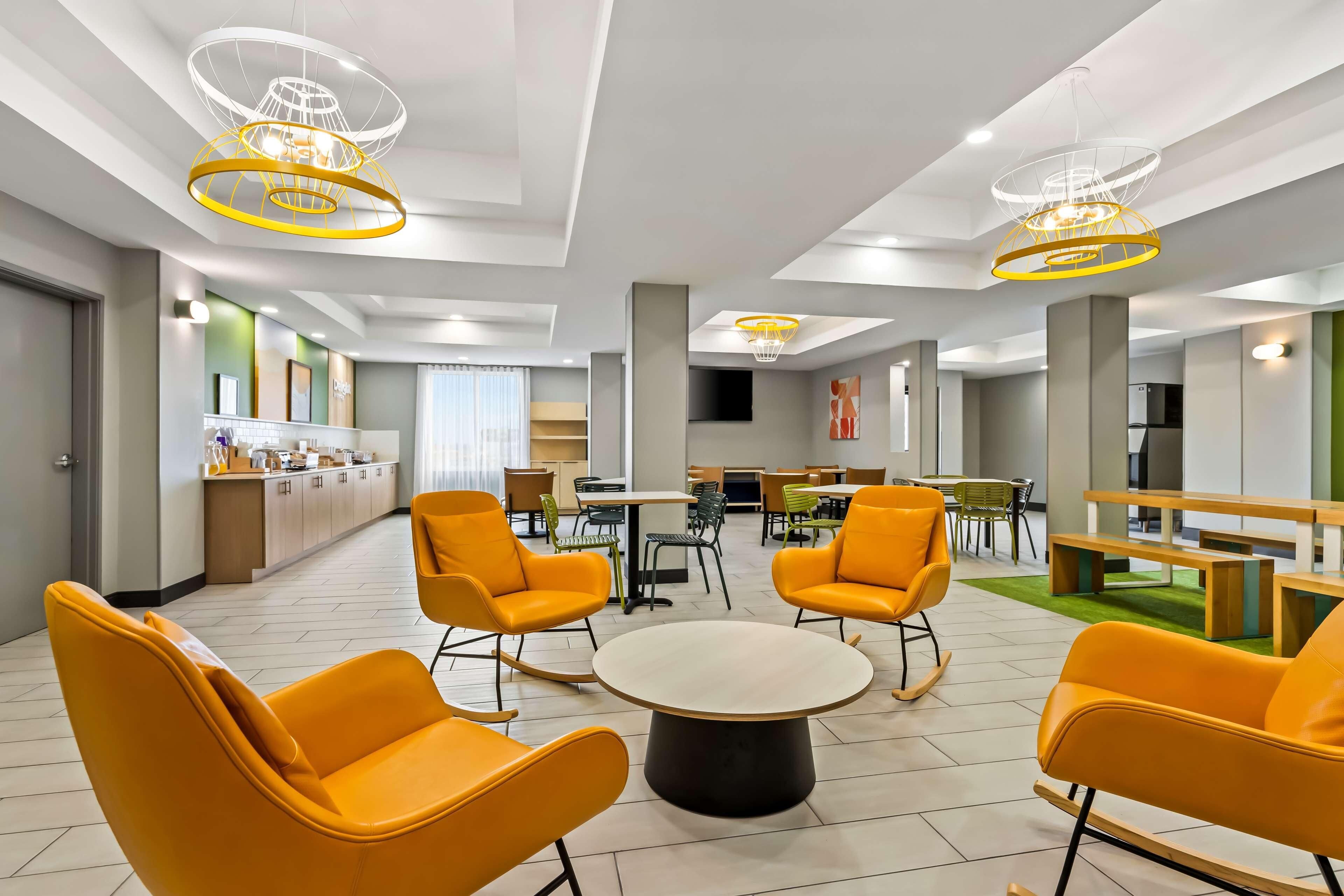 A modern hotel lobby with bright orange chairs, a central round table, and unique ceiling lights.