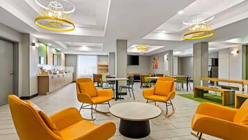 A modern hotel lobby with bright orange chairs, a central round table, and unique ceiling lights.