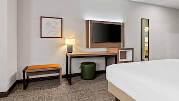 Quarto de hotel com uma cama, escrivaninha com cadeira, televisão, abajur, quadro emoldurado e lixeira.