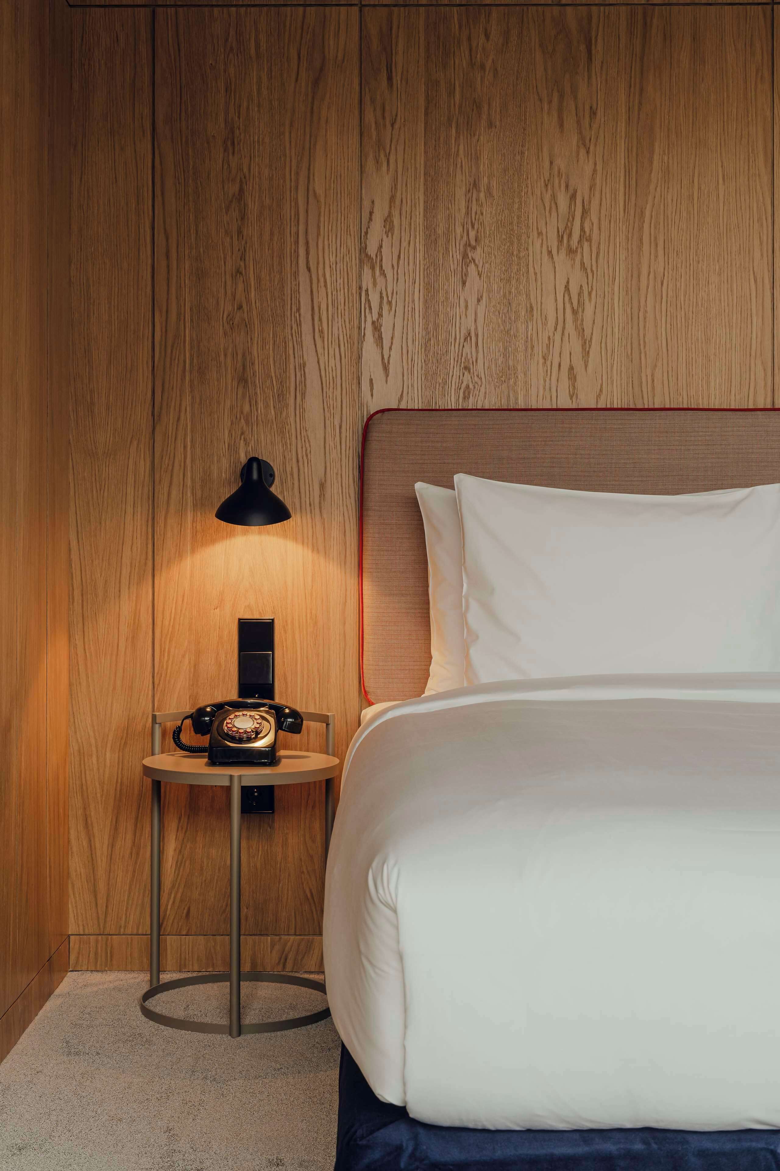 Una cama bien hecha con sábanas blancas, un cabecero de madera, una mesita de noche con un teléfono antiguo y una lámpara negra.