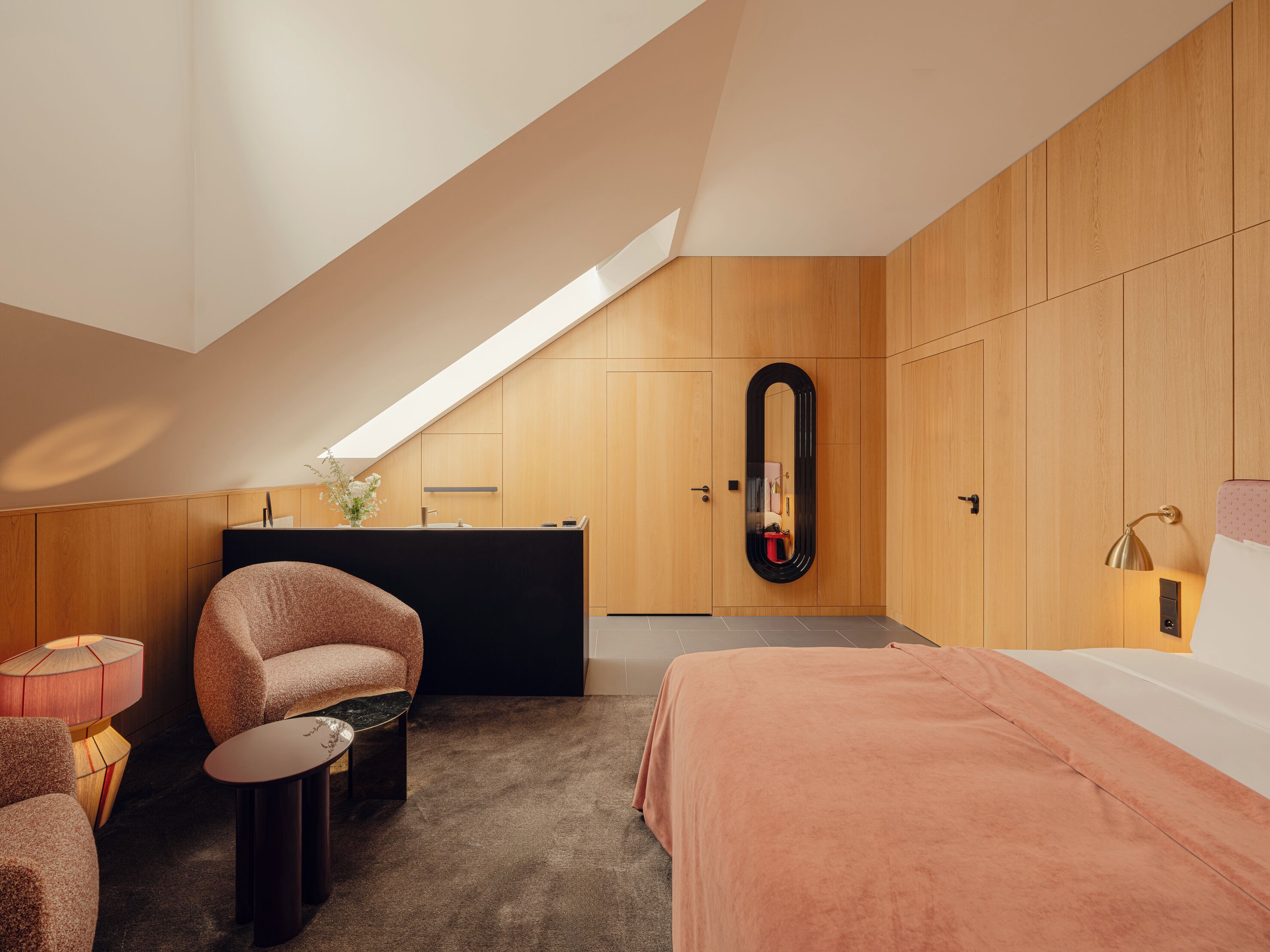 Un dormitorio moderno con techo inclinado, un escritorio empotrado, un espejo grande, una cama y una pequeña zona de estar.