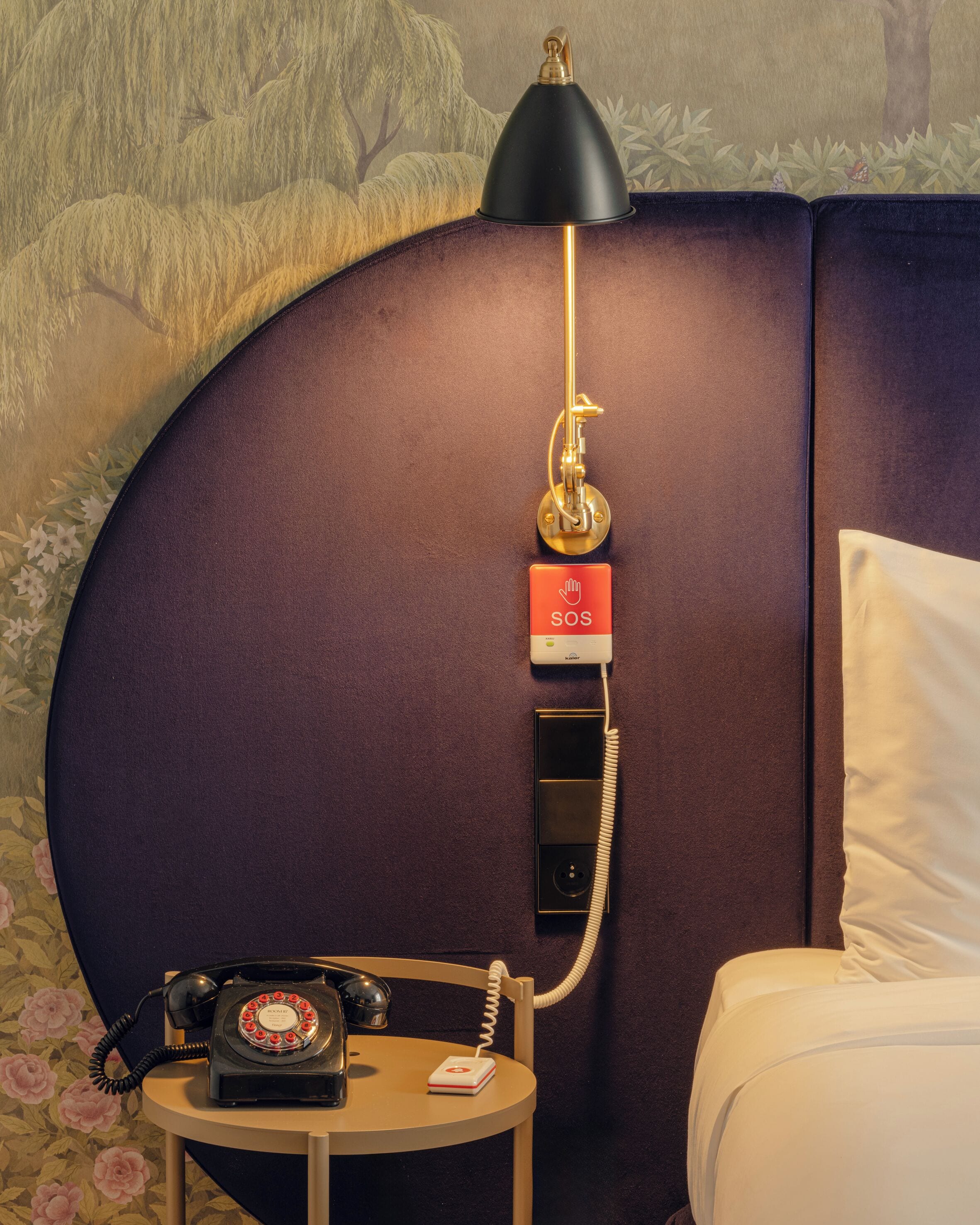 Camera d'albergo con un letto, un comodino con telefono a disco, un telefono di emergenza SOS montato a parete e una lampada.