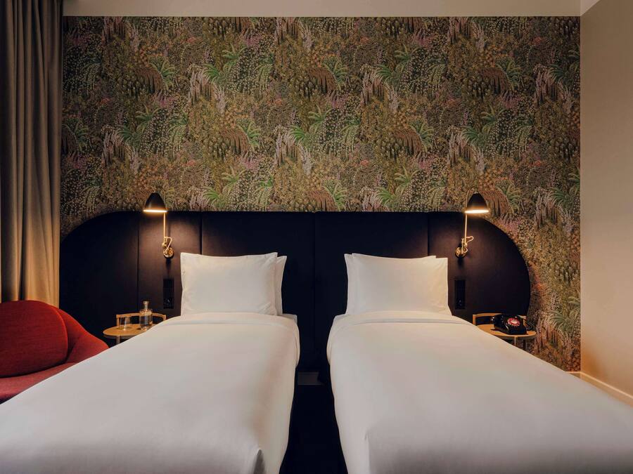Ein Hotelzimmer mit zwei Betten, einem roten Sessel und einer Wand mit einem dichten Dschungelmotiv.