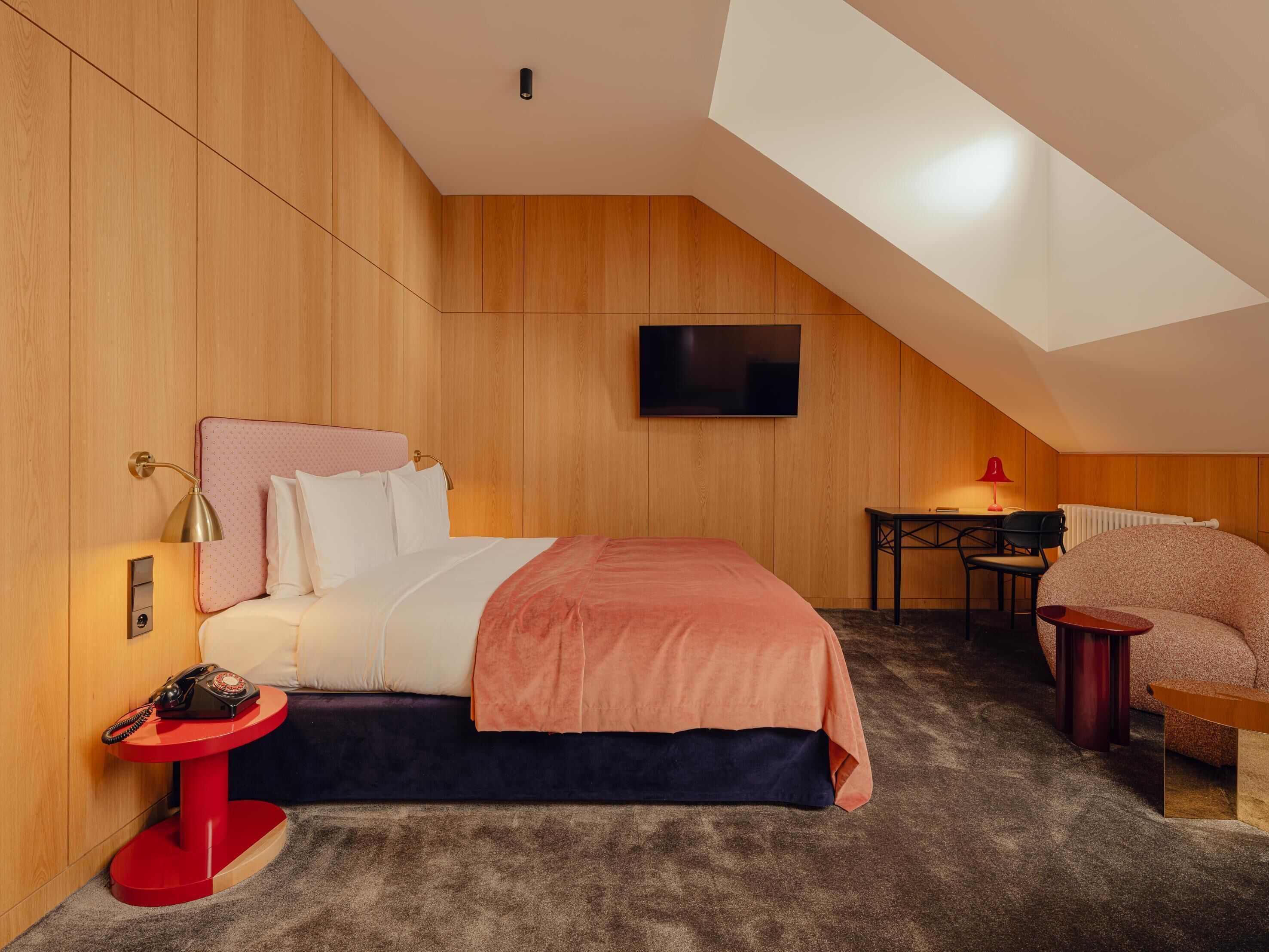 Un dormitorio moderno con techo inclinado, una cama con cabecera rosa, una mesita de noche roja, un escritorio con silla y un sillón redondo.