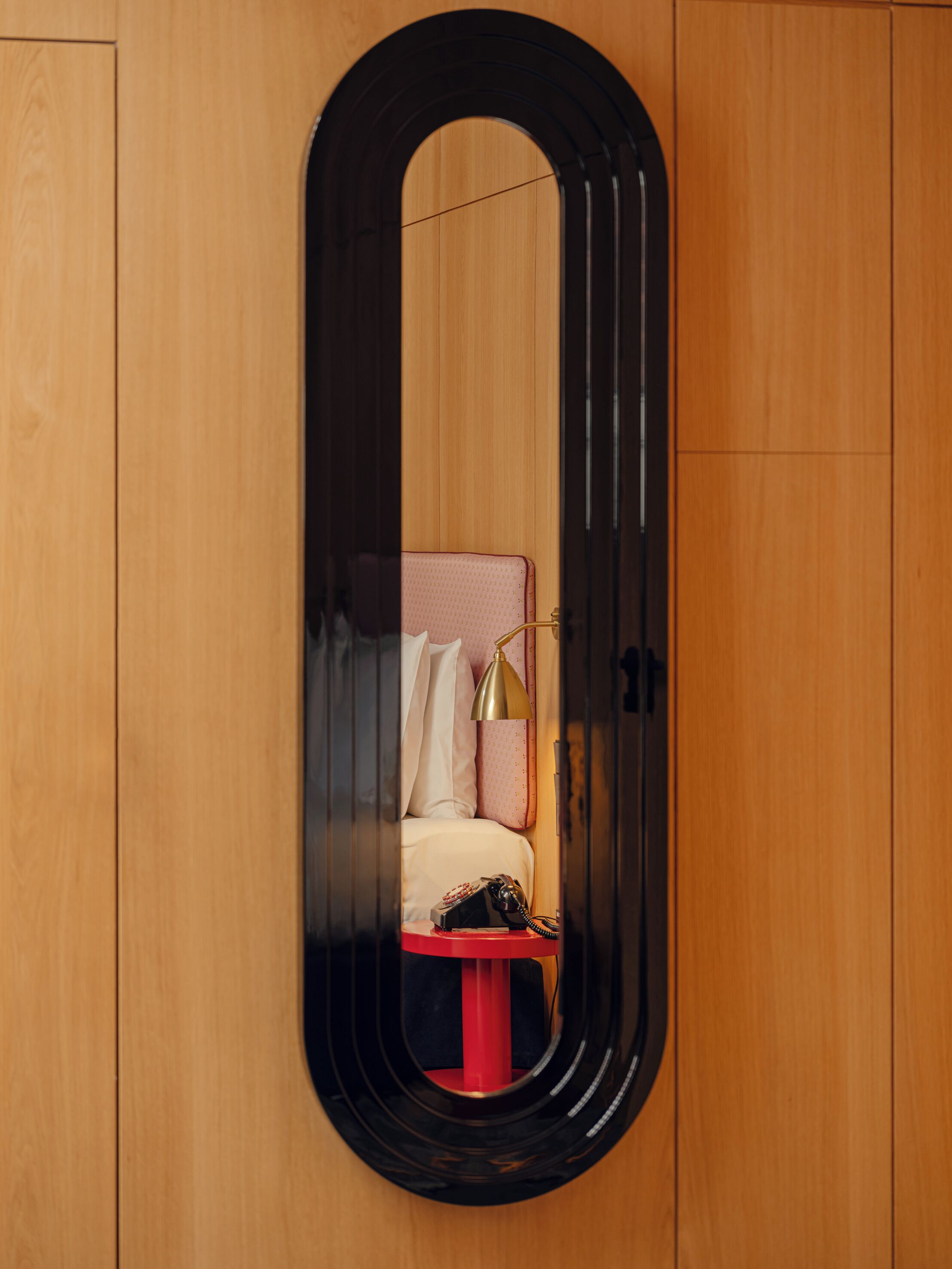 A bedroom with a wooden wardrobe, a bed with a red nightstand, and a mirror with a black frame.