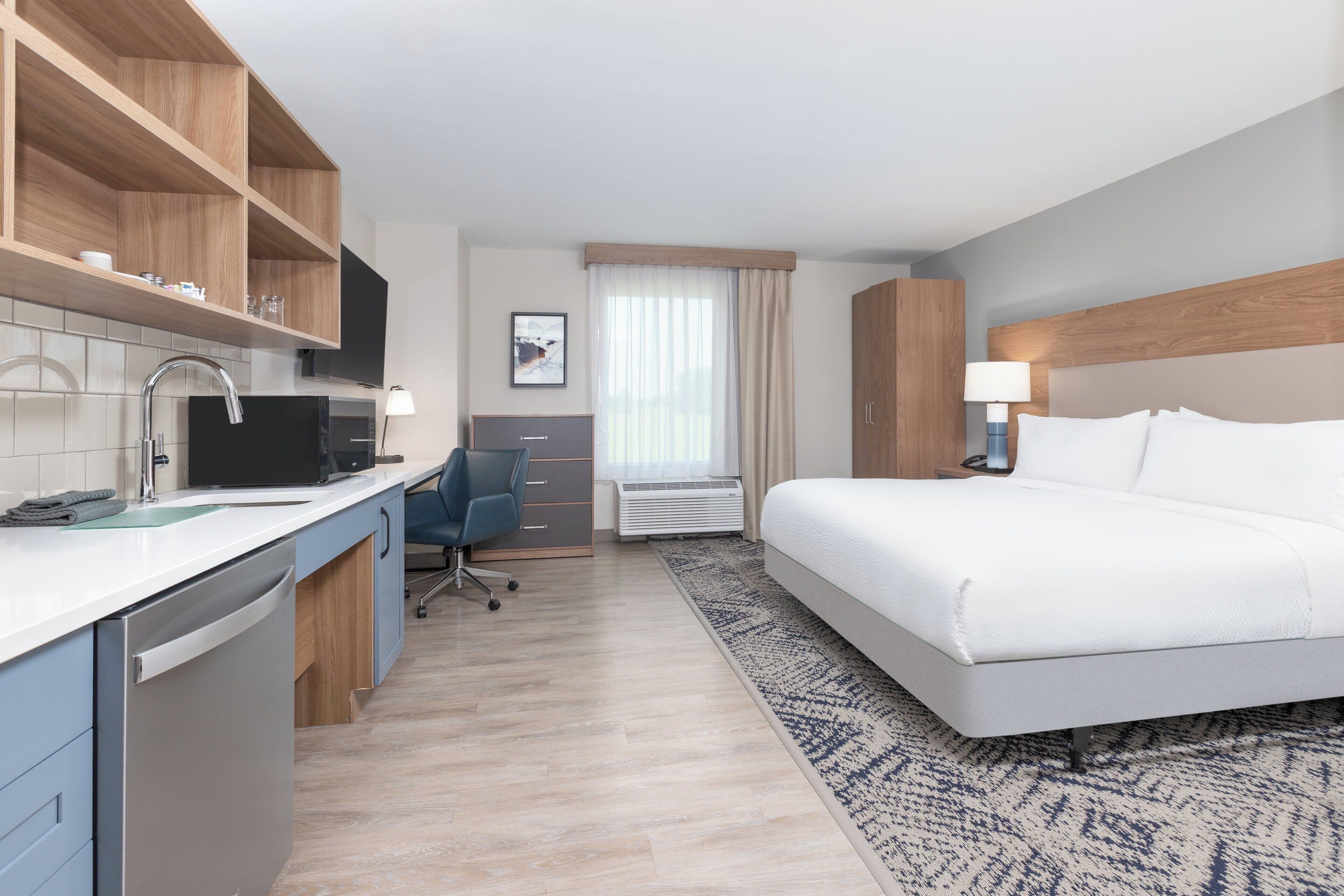 A hotel room with a bed, a desk with a chair, a kitchen area with a dishwasher, and a window with curtains.