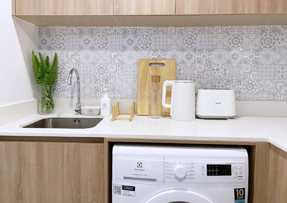 A kitchen with a sink, a washing machine, a cutting board, a mug, and a plant.