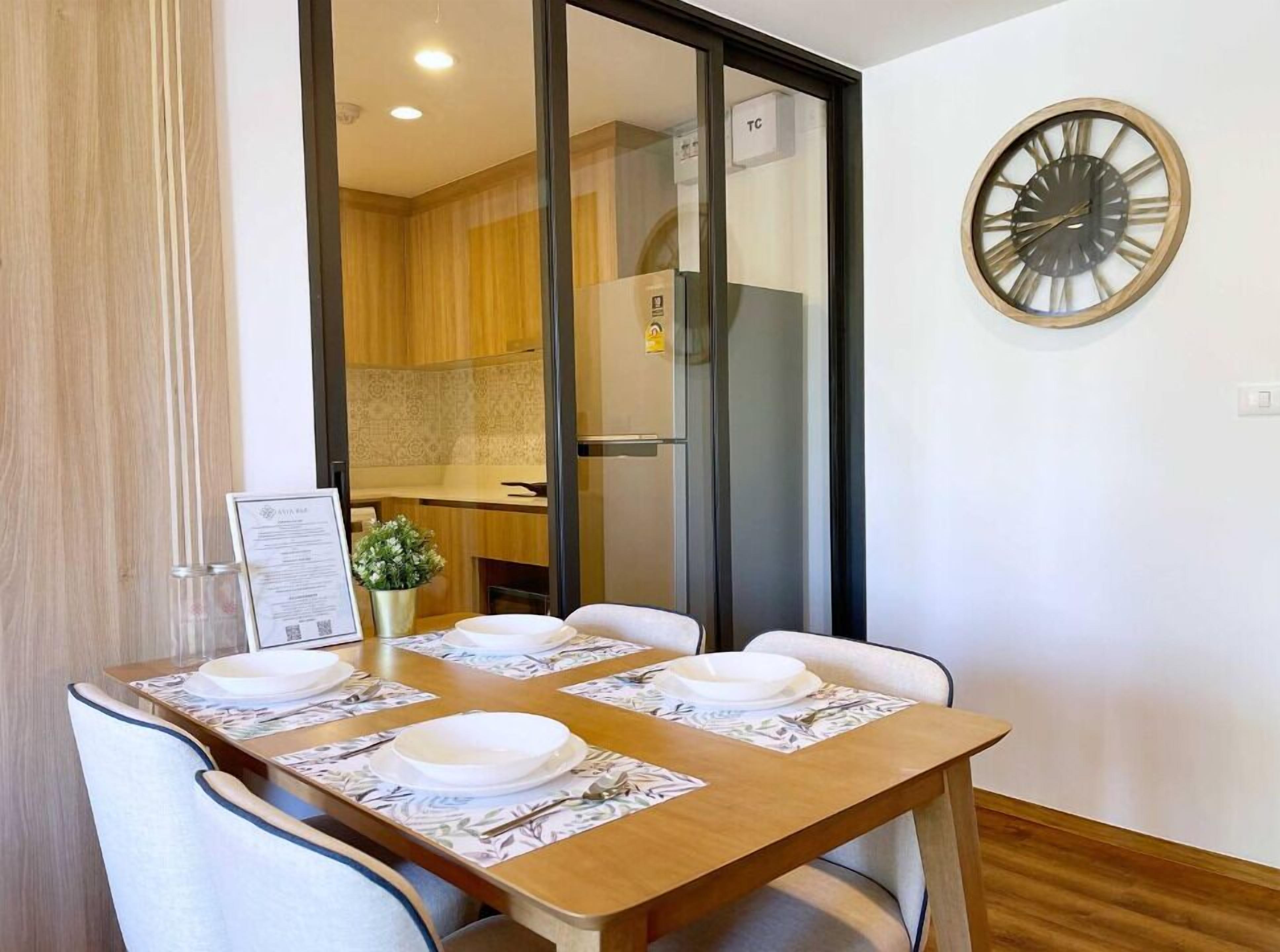 Uma sala de jantar com uma mesa de madeira posta para quatro pessoas, um relógio na parede e vista para a cozinha.