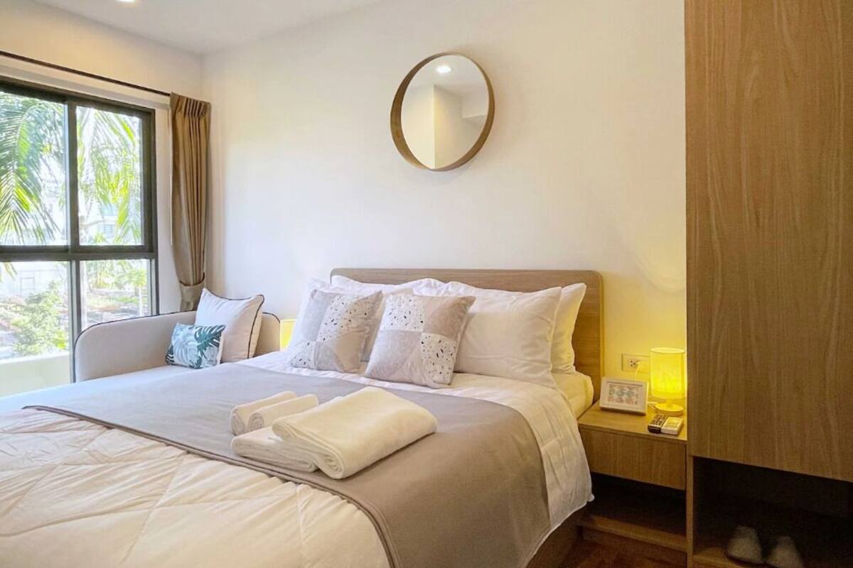 Una cama bien hecha con sábanas blancas y una toalla doblada, una mesita de noche con una lámpara y un espejo en la pared.