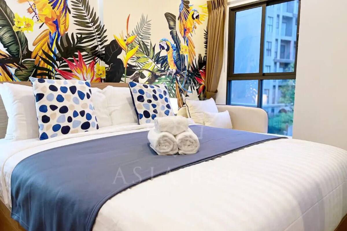 Una cama bien hecha con cubrecama azul y almohadas blancas, un mural temático tropical y una ventana con vistas a edificios.
