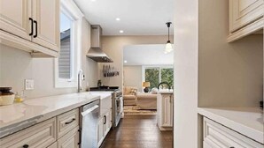 A modern kitchen with white cabinetry, a central island, and a view to a living area. - Perfection Near the River (Niagara Falls)