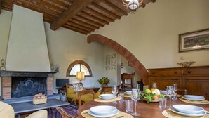 A dining area with a wooden table set for four, surrounded by chairs. A fireplace with a mantel is visible in the background.