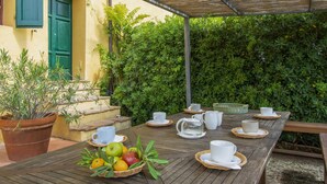 An outdoor dining area with a wooden table set for tea, surrounded by lush greenery and a yellow building with green shutters.