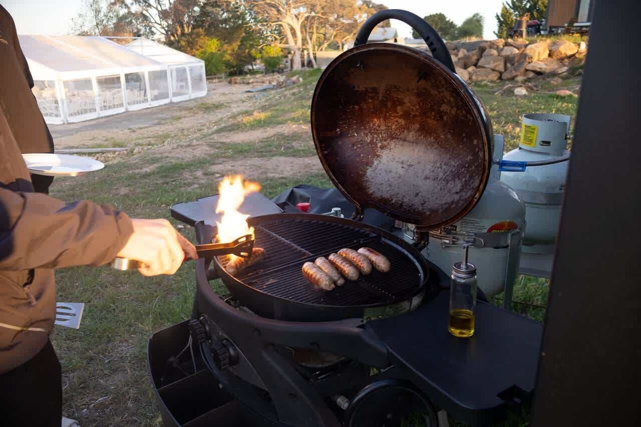 A portable grill with sausages being cooked, a bottle of oil, and a gas canister.