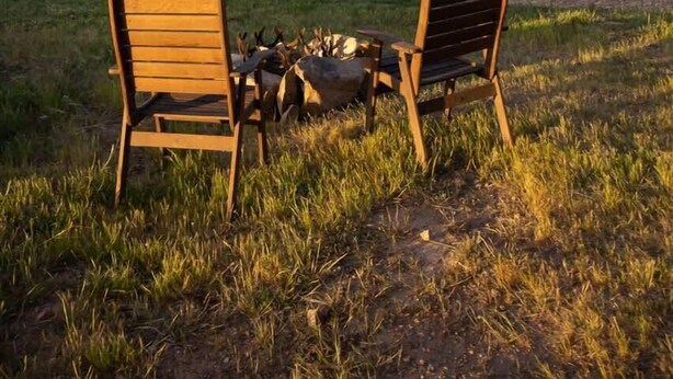 Twee houten stoelen gericht op een vuurkuil in een grasveld, met een boom op de achtergrond.