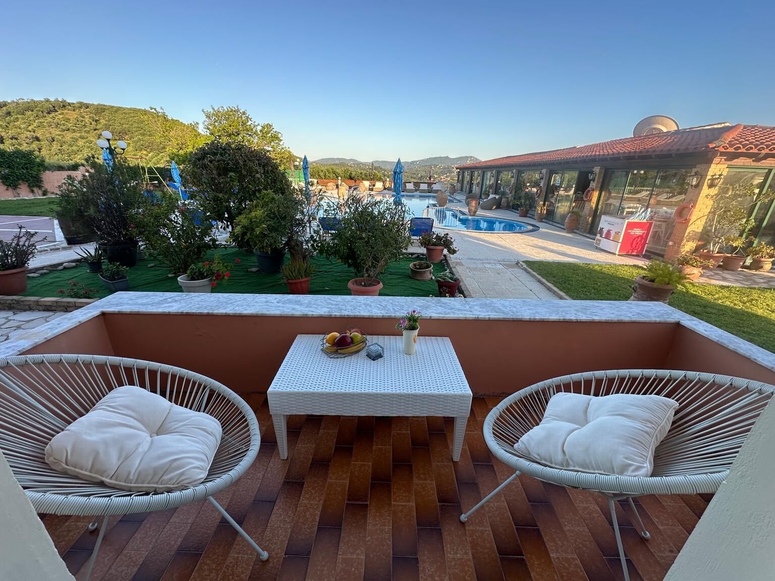 Ein Balkon mit Korbstühlen und einem Tisch, mit Blick auf einen Pool und viel Grün.