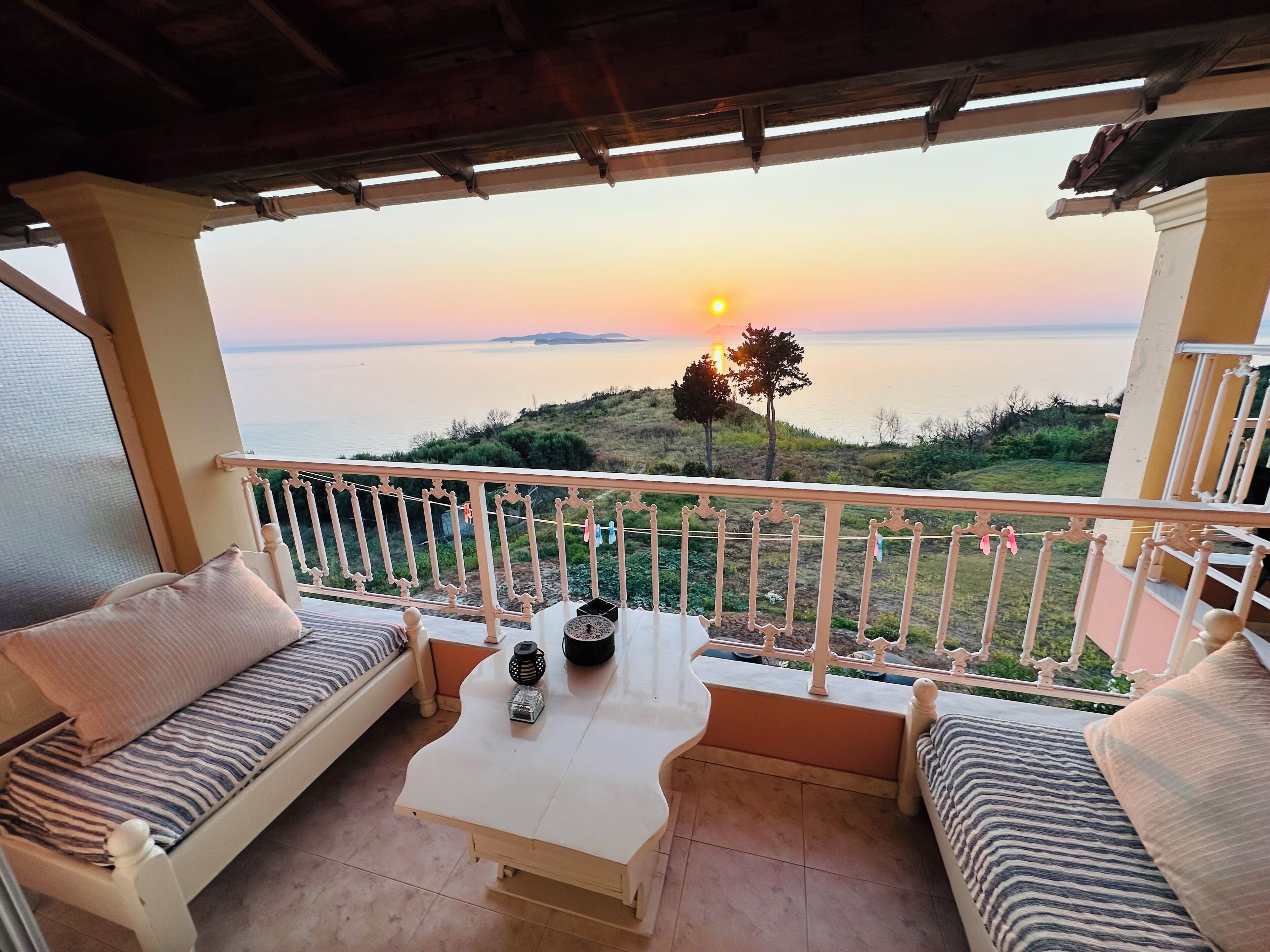 Ein Balkon mit einem weißen Tisch, zwei Sitzgelegenheiten mit Kissen und Blick auf den Sonnenuntergang über dem Meer.