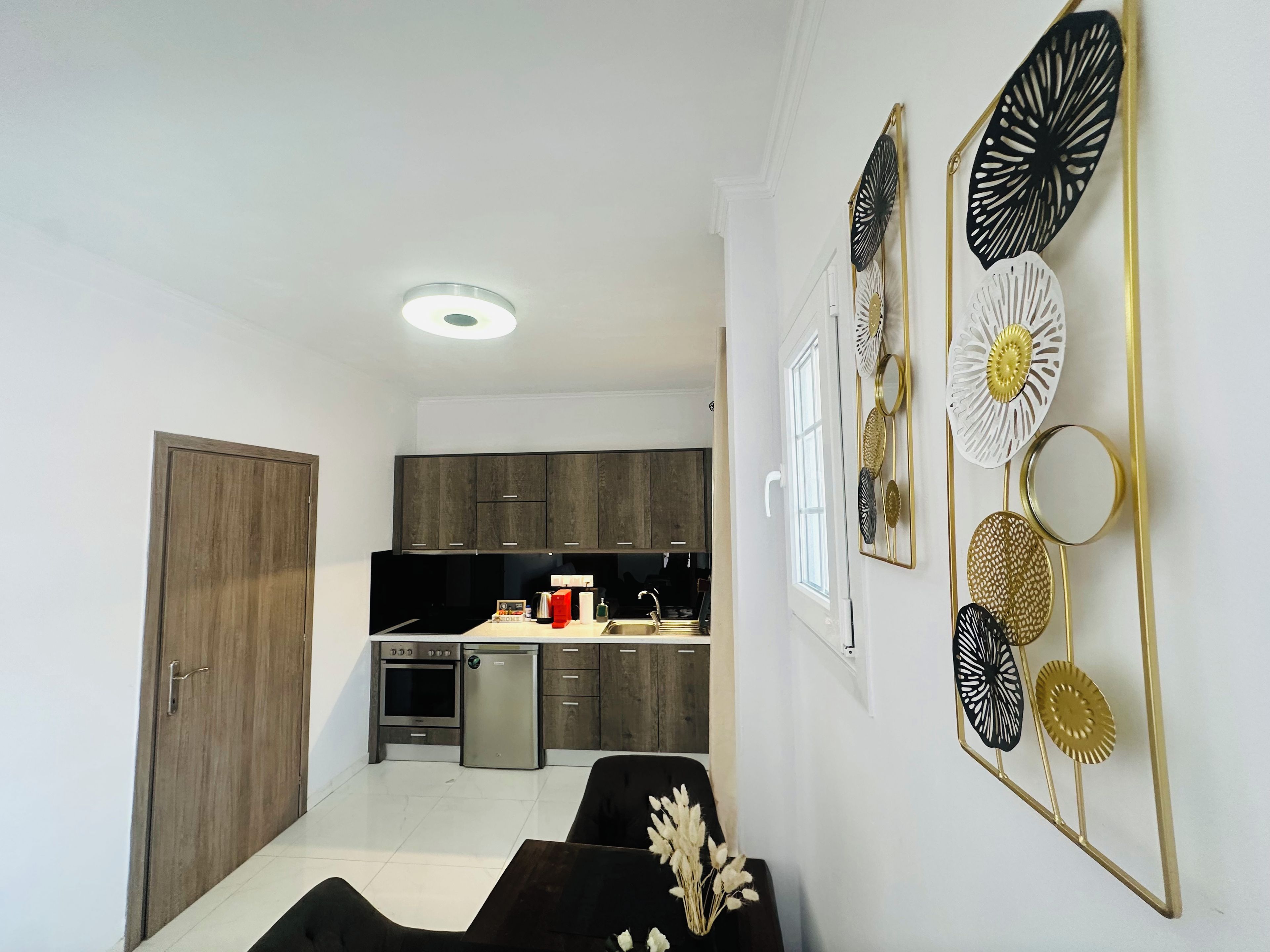 Une cuisine moderne avec des meubles en bois foncé, un four encastré et un réfrigérateur. On y trouve un coin repas avec une table et des chaises noires, ainsi qu’un élément décoratif mural orné de motifs circulaires et floraux.