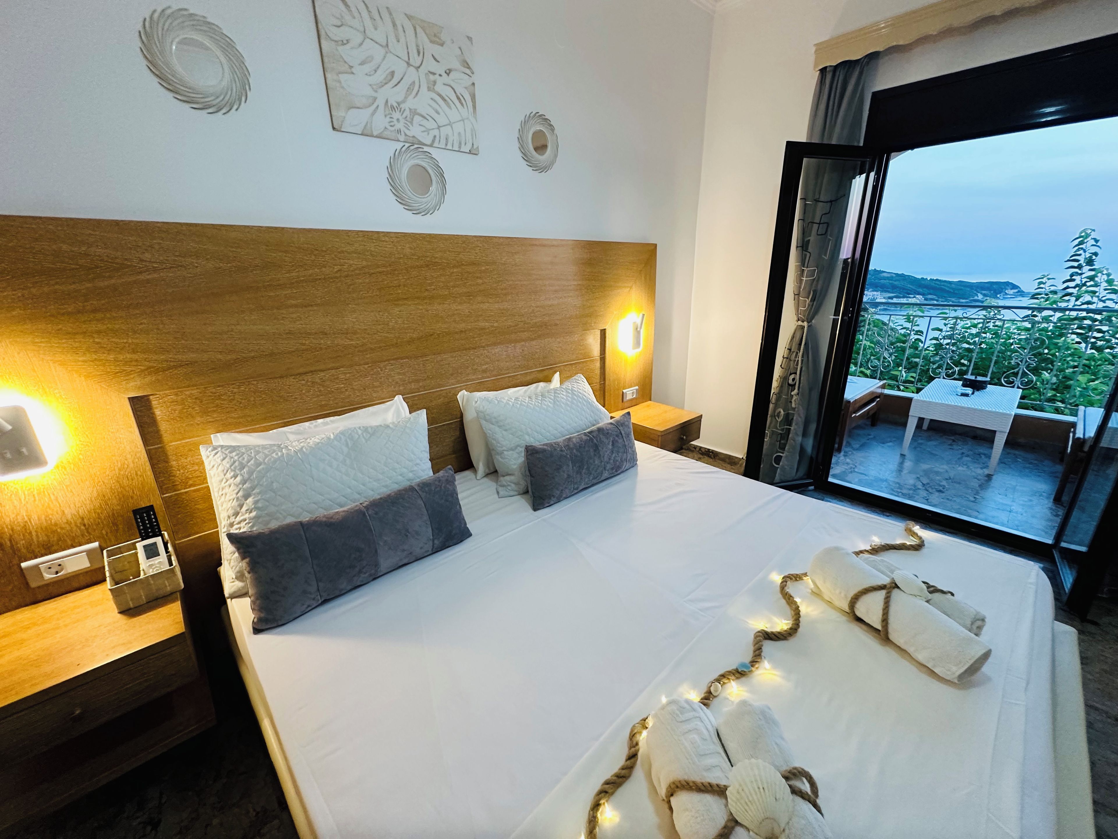 A hotel room with a large bed, two bedside lamps, a wooden headboard, and a view of a balcony with outdoor furniture.