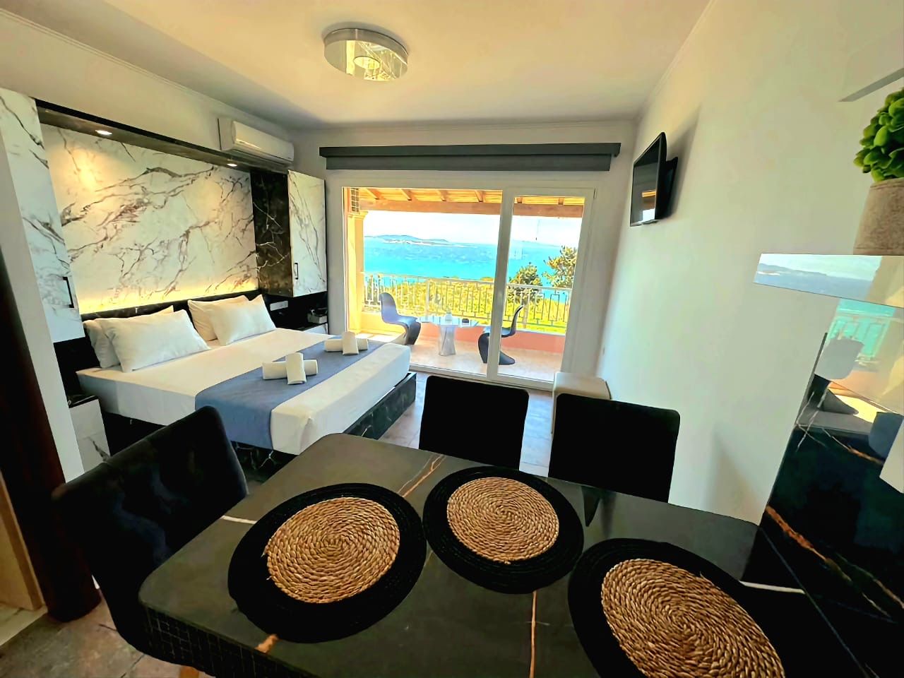 Quarto de hotel moderno com cama, área de jantar e varanda com vista.