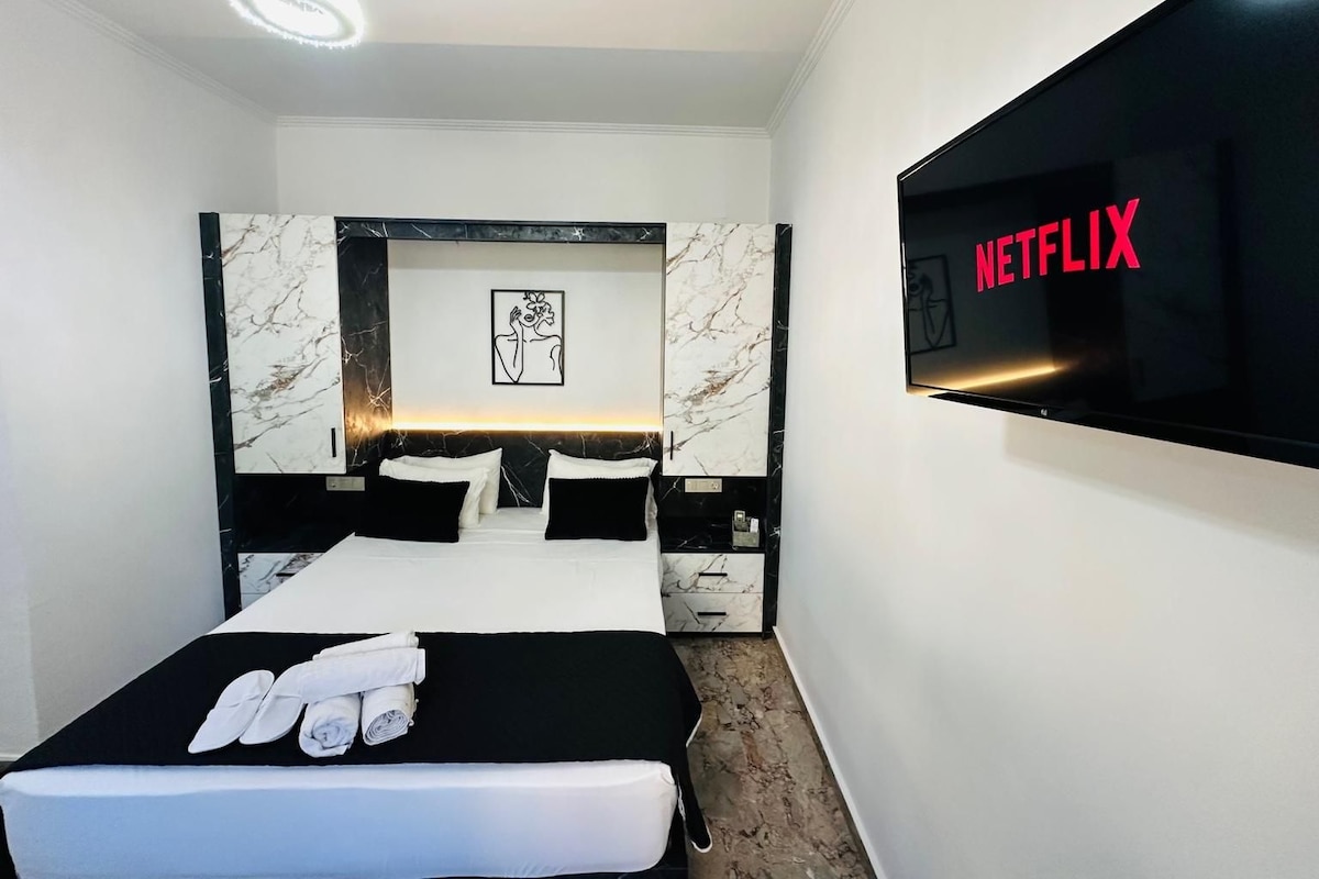 Una habitación de hotel moderna con una cama grande, un televisor de pantalla plana mostrando el logo de Netflix, cabecera con diseño de mármol y una obra de arte enmarcada sobre la cama.
