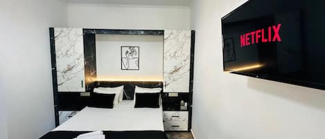 Une chambre d’hôtel moderne avec un grand lit, un téléviseur à écran plat affichant le logo de Netflix, une tête de lit à motifs de marbre et une œuvre d’art encadrée au-dessus du lit.