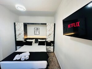 A modern hotel room with a large bed, a flat-screen TV displaying the Netflix logo, marble-patterned headboard, and a framed artwork above the bed.