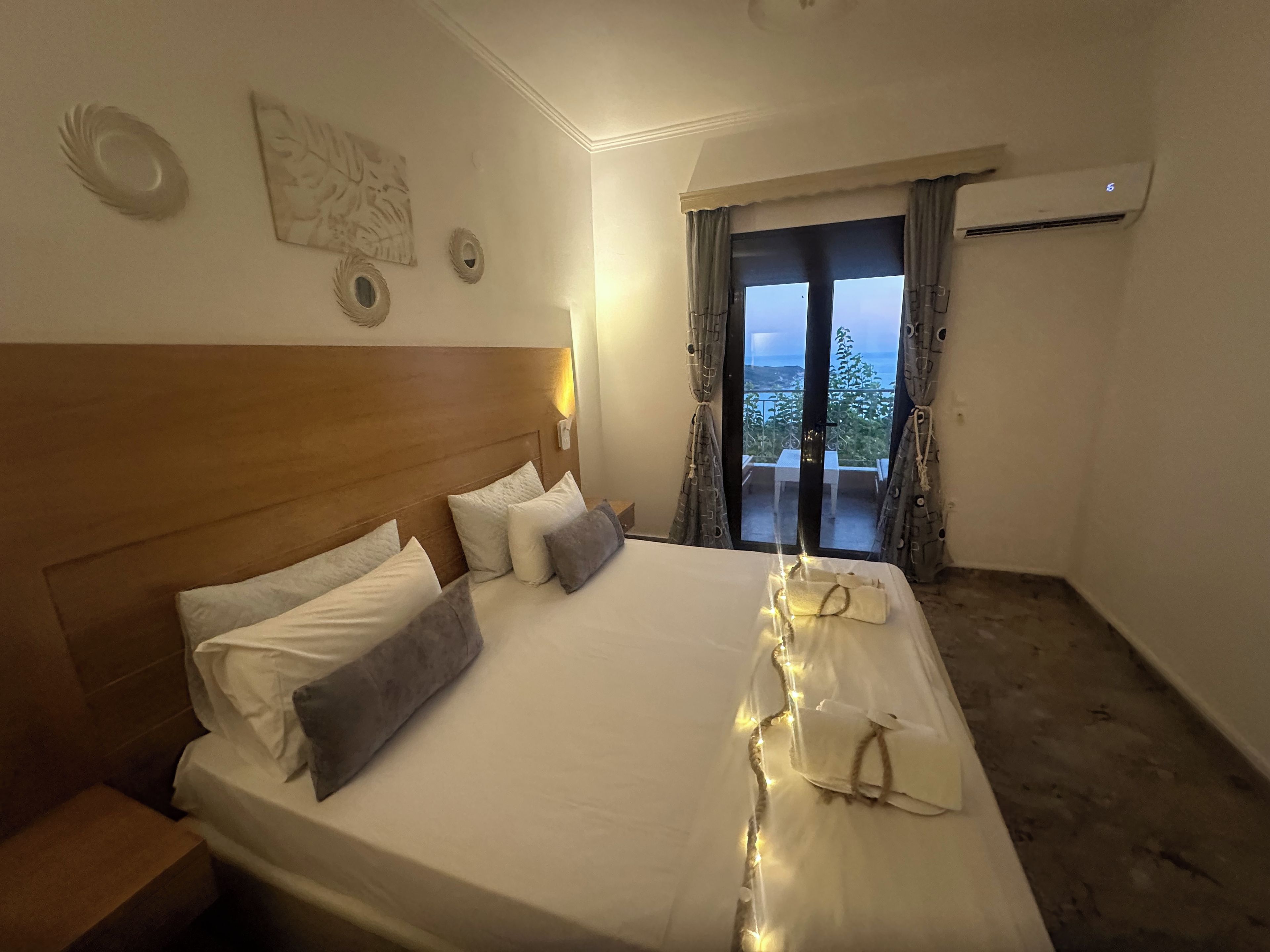 Ein Hotelzimmer mit einem großen Bett, einem Nachttisch, einer Wand-Klimaanlage und Blick ins Freie durch ein Fenster mit Vorhängen.